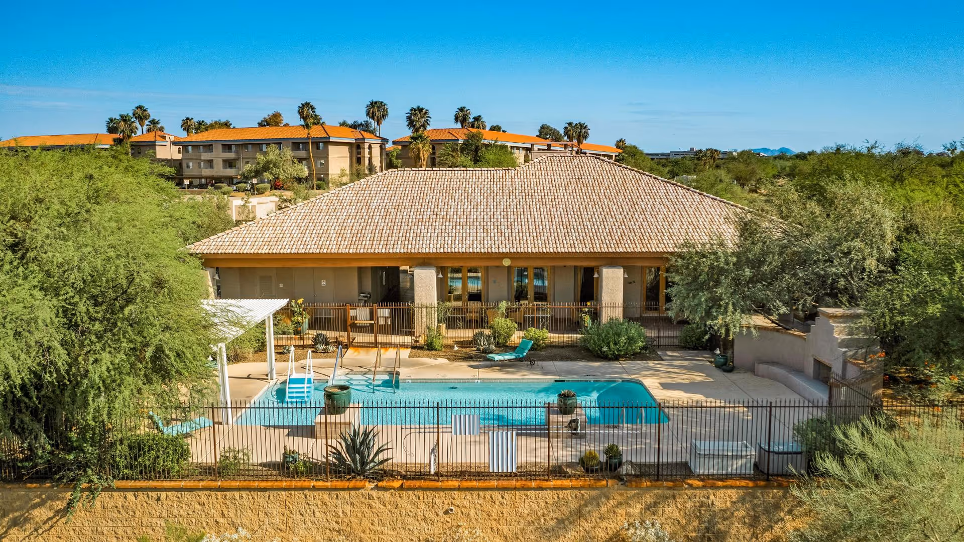 Outdoor pool and patio in front of a single-story clubhouse with a tiled roof, surrounded by desert landscaping and nearby apartment buildings.