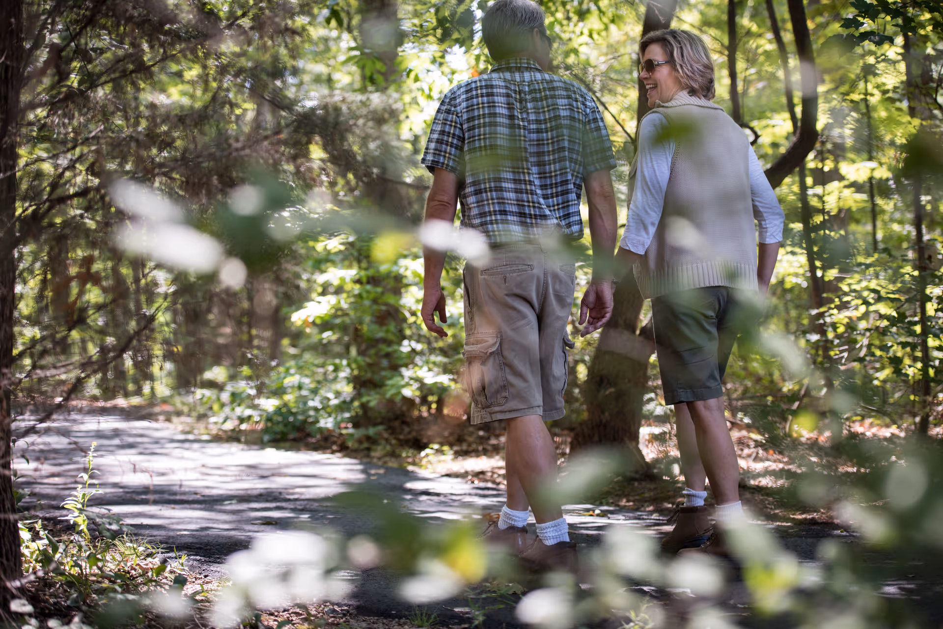 An older couple walking hand in hand on a shaded forest path surrounded by trees and greenery, enjoying a leisurely outdoor stroll.