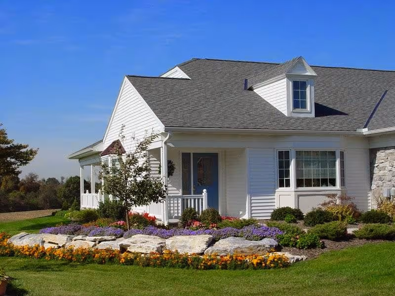A white single-story house with a gray shingled roof, a small front porch with a blue door, and a well-maintained garden featuring colorful flowers and shrubs in front of the house under a clear blue sky.