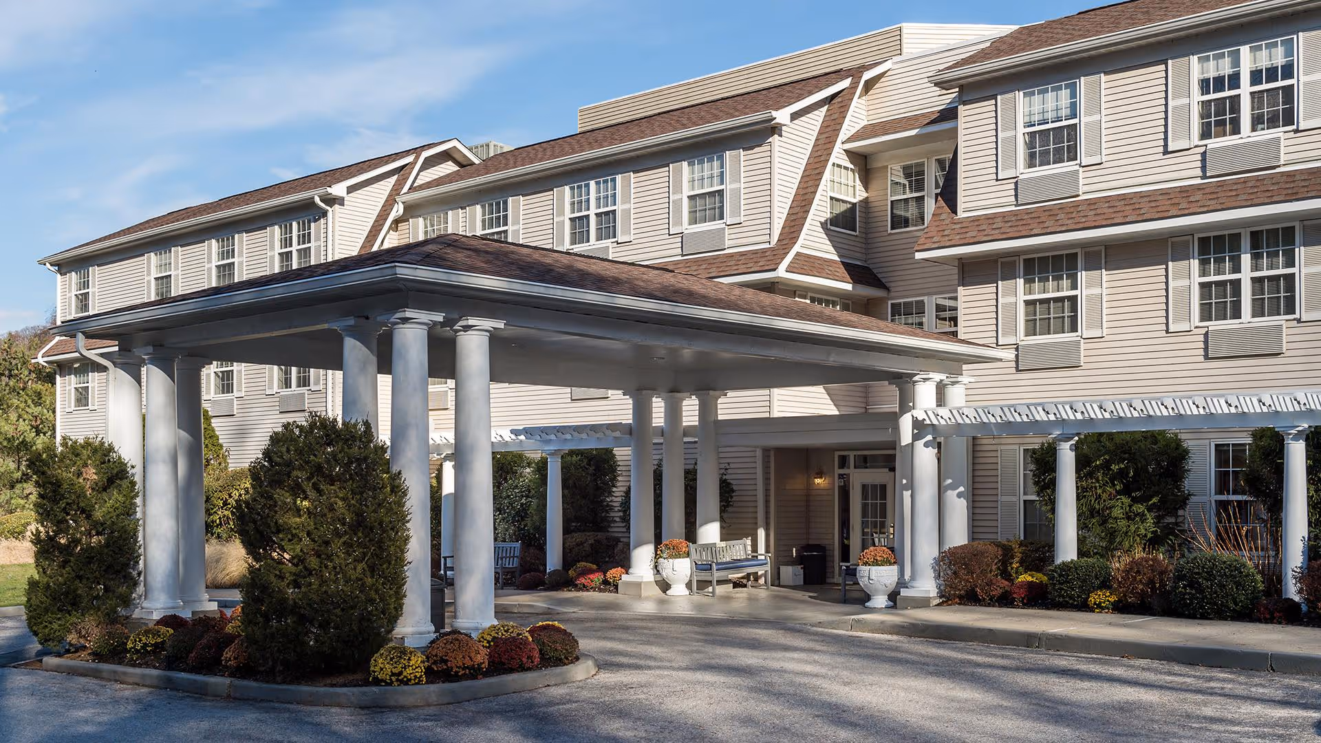 Front entrance porte-cochère of a multi-story senior living building with white columns, landscaping, and benches.