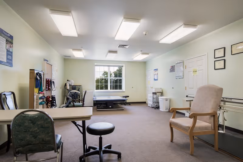 Well-lit interior therapy/rehab room with chairs, tables, exercise equipment, and a treatment table by a window.
