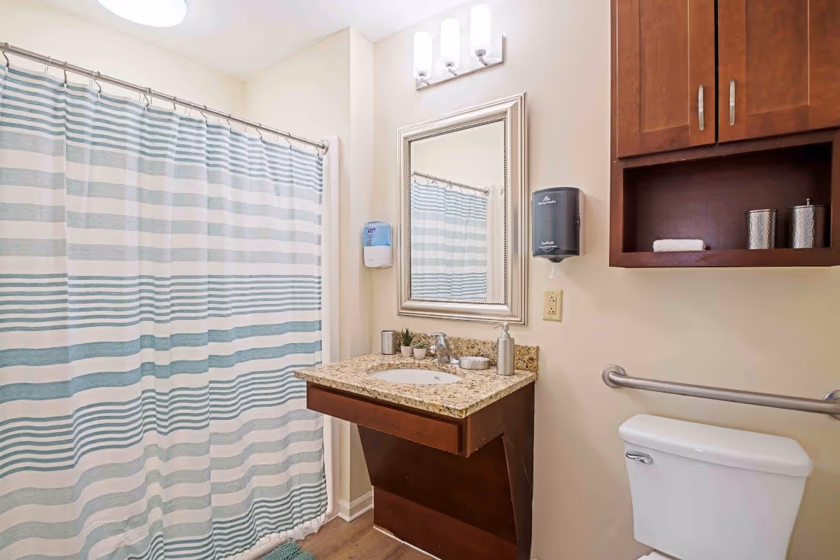 A clean and well-lit bathroom featuring a shower with a striped curtain, a granite countertop with a sink, a mirror above the sink, a wall-mounted soap dispenser, a wooden cabinet with towels and containers, a toilet with a grab bar, and light-colored walls.