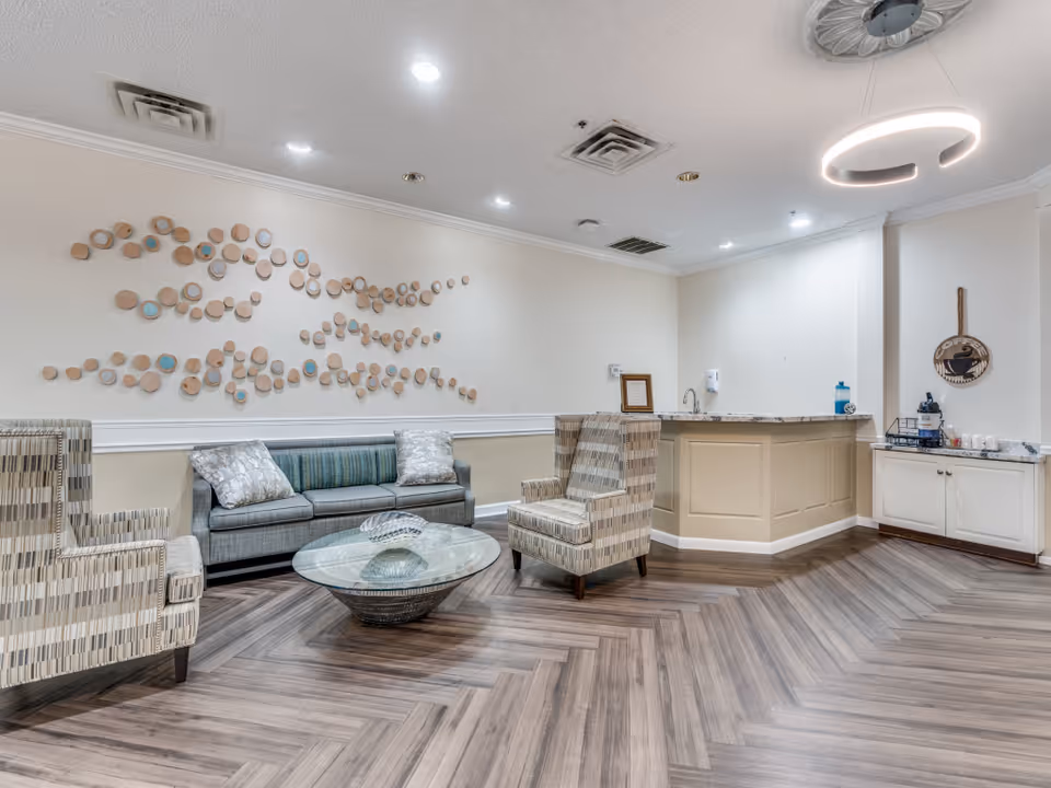 A modern lounge area with a gray sofa and two patterned armchairs around a glass coffee table. The room features a decorative wall installation of circular wooden pieces, a small counter with a sink, and a coffee station with cups and a coffee sign on the wall. The floor has a herringbone wood pattern and the ceiling has recessed lighting and a circular hanging light fixture.