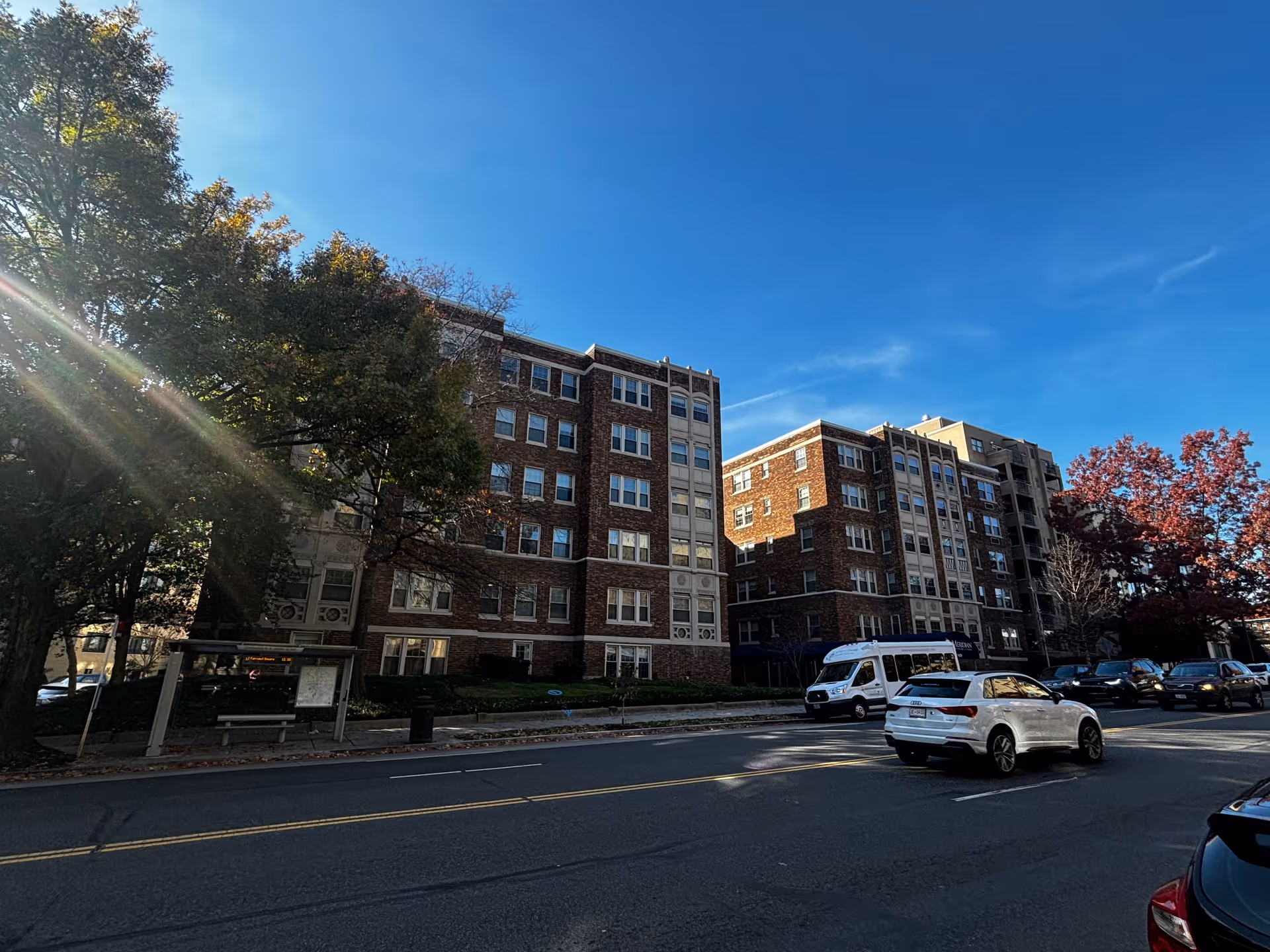 Street view of a multi-story brick building with multiple windows, surrounded by trees with autumn foliage. Several cars are parked and driving on the road in front of the building under a clear blue sky.