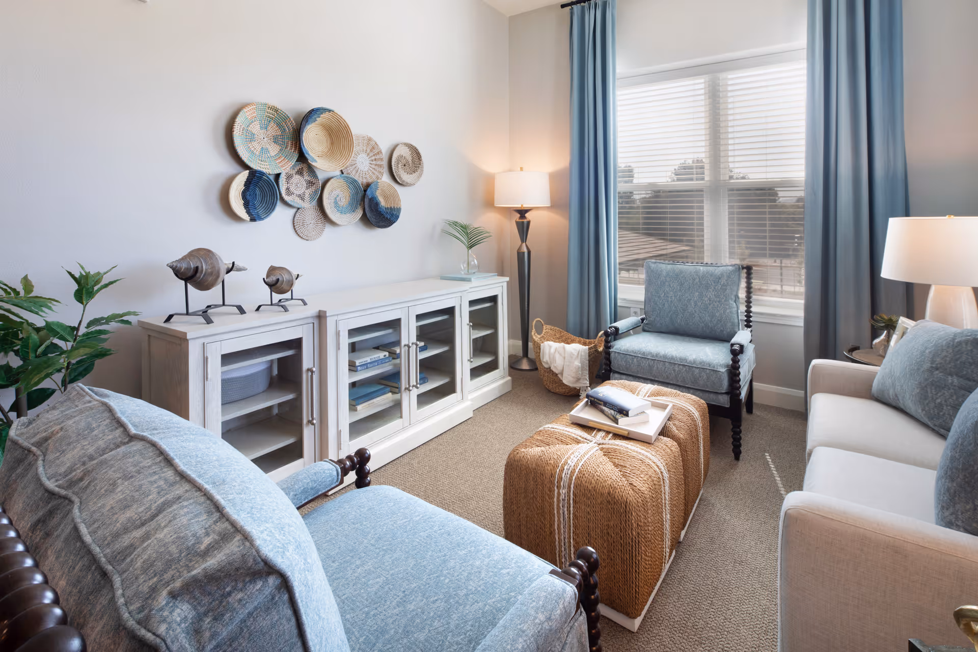 A cozy living room with light blue upholstered armchairs and a sofa, a woven ottoman coffee table with books on top, a white cabinet with glass doors, decorative wall baskets, a floor lamp, and large windows with blue curtains letting in natural light.