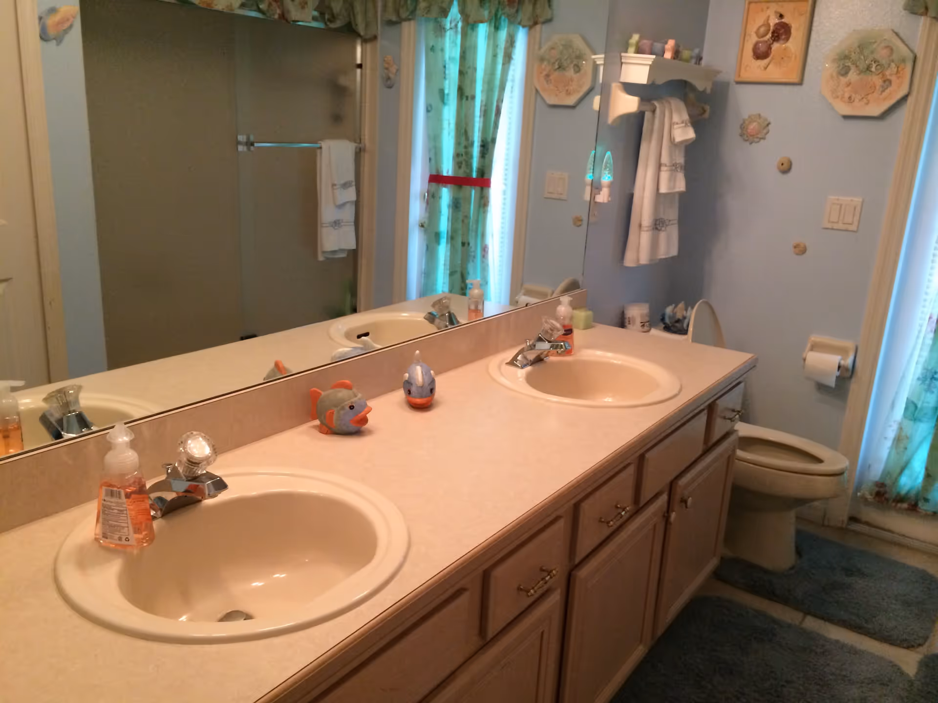 A bathroom with a double sink vanity featuring two beige sinks and a large mirror above. On the countertop are two fish-shaped soap holders and a bottle of liquid soap. The bathroom has light blue walls decorated with framed artwork and small wall hangings. A toilet is visible next to the vanity, along with a toilet paper holder and a blue bath mat on the floor. A shower with a frosted glass door and a towel hanging on the bar is seen in the background. The window is covered with floral curtains.