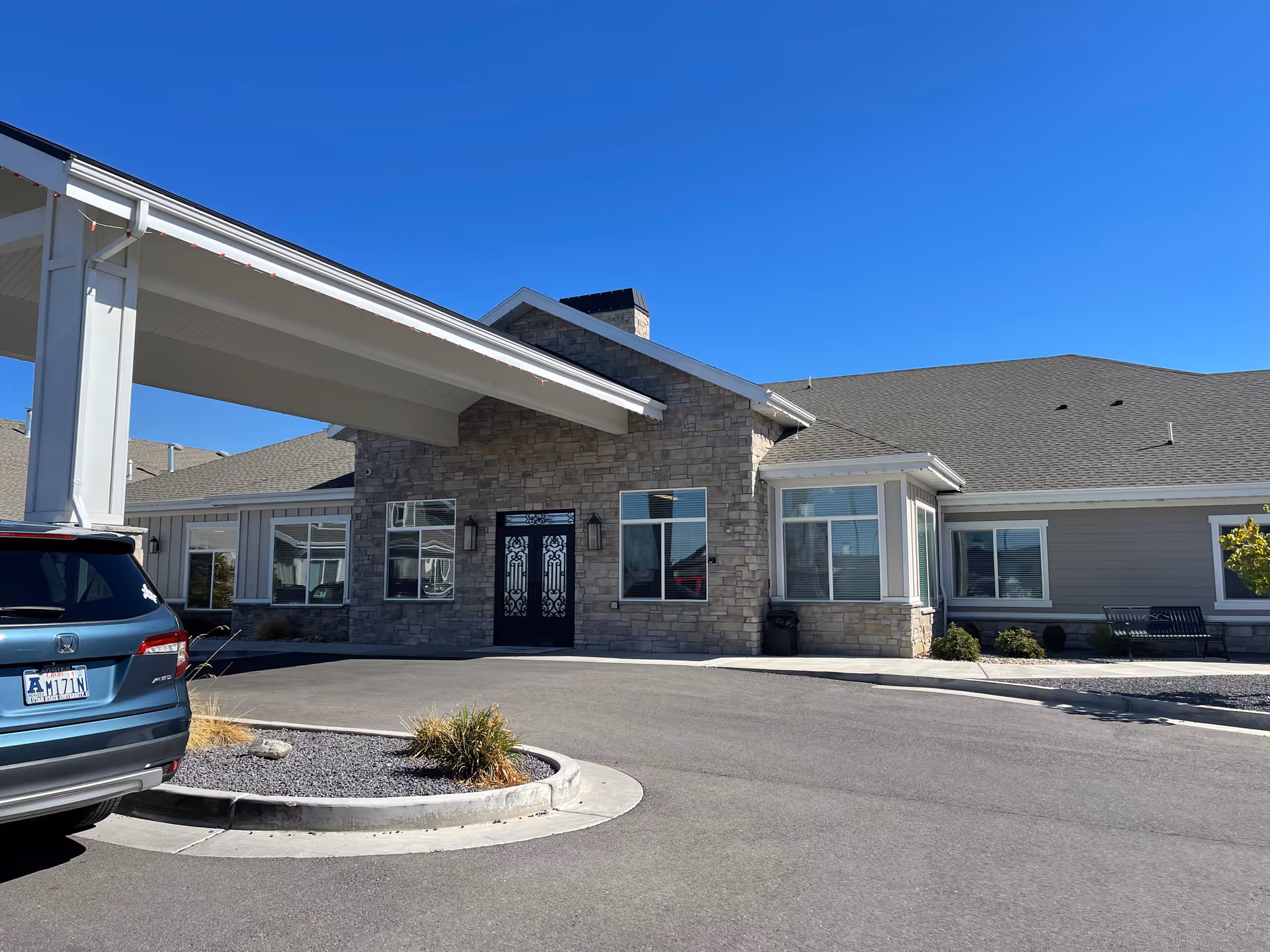 Exterior view of Sunridge Assisted Living of Layton showing the main entrance with stone facade, large windows, and a covered drop-off area. A blue SUV is parked nearby under a clear blue sky.