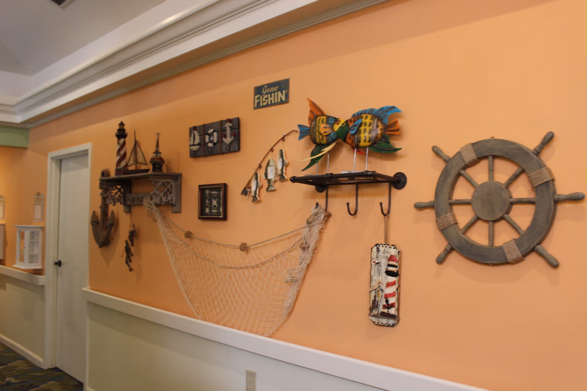 A hallway with peach-colored walls decorated with nautical-themed items including a ship's wheel, fishing net, model sailboats, a lighthouse, colorful fish sculptures, and signs with maritime motifs.
