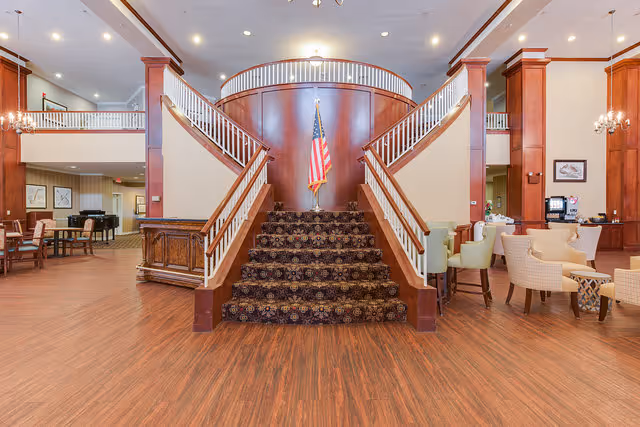 Spacious interior of a senior living facility featuring a central carpeted staircase with wooden railings leading to an upper level. An American flag is displayed at the top of the stairs. The room has wood flooring, wooden columns, and various seating areas with chairs and tables on either side. The ceiling is equipped with recessed lighting and chandeliers.