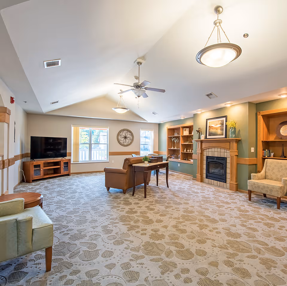 A spacious and well-lit living room with a patterned carpet, a ceiling fan, and two hanging light fixtures. The room features a fireplace with a wooden mantel, built-in shelves with decorative items, a flat-screen TV on a wooden stand, a brown leather sofa, a wooden table, and two armchairs. Large windows allow natural light to fill the space.