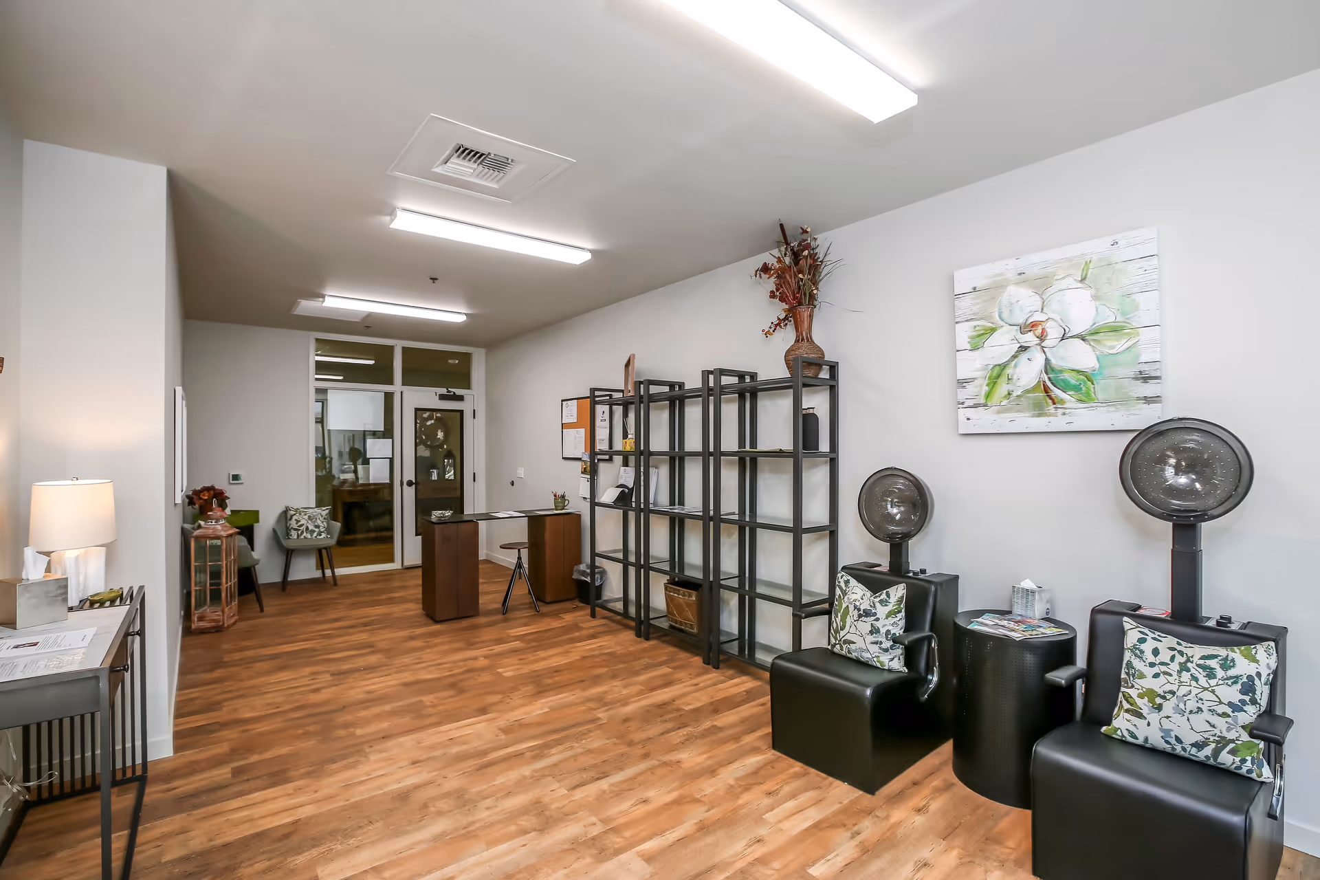 Interior room with wooden flooring and white walls featuring two black salon hair dryer chairs with floral cushions, a small round black table between them, a black metal shelving unit with various items, a wooden desk with a chair, and a small sitting area with a chair and side table. A floral painting hangs on the wall above the hair dryer chairs.