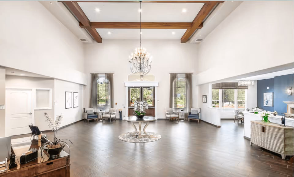 Spacious senior living facility lobby with high ceilings, wooden beams, a large chandelier, and a round table with a flower arrangement in the center. There are multiple seating areas with chairs near tall windows with sheer curtains, and a reception desk area on the right side.