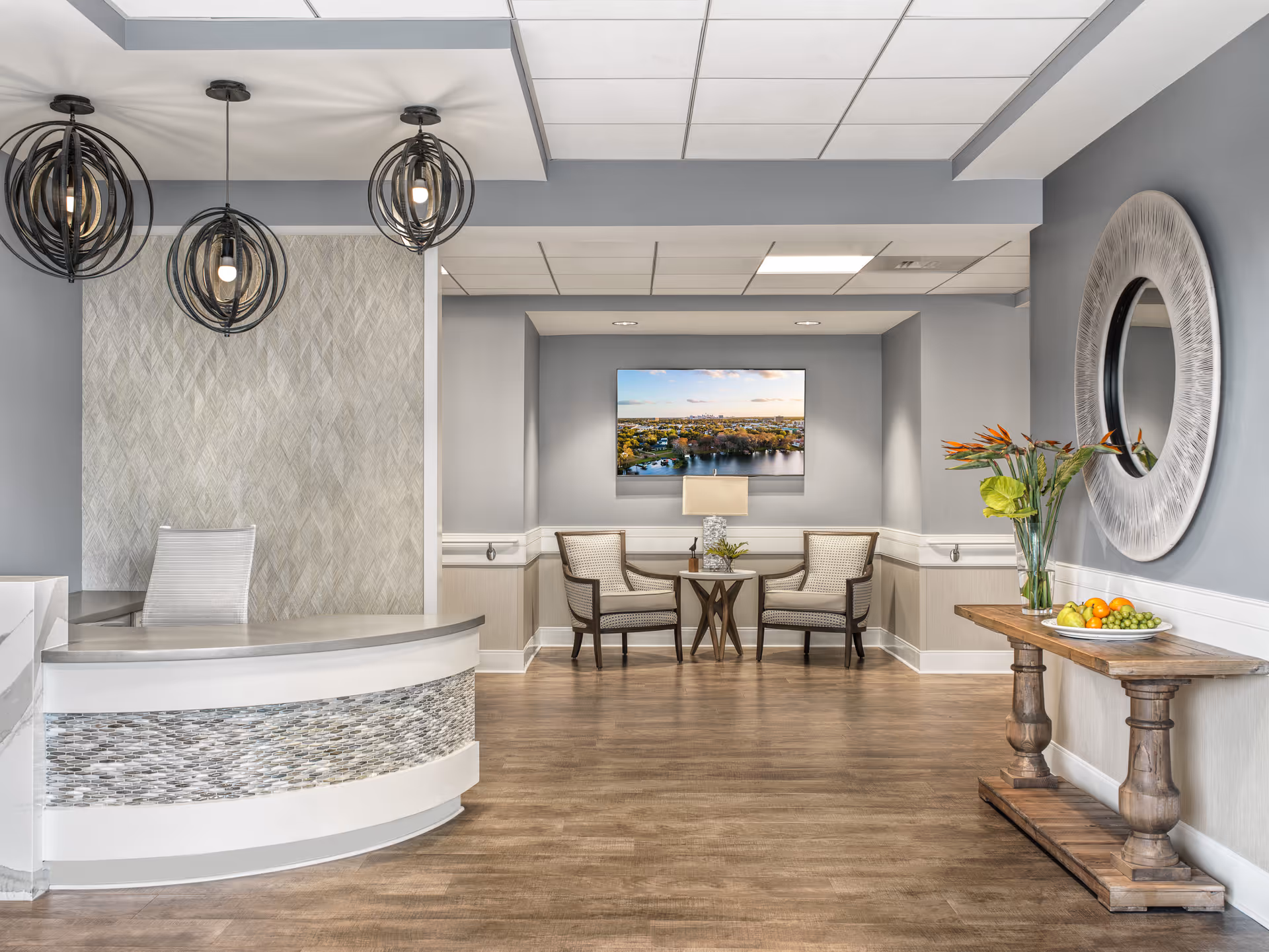 A modern reception area with a curved desk featuring a mosaic tile front, three spherical pendant lights hanging from the ceiling, and a seating area with two upholstered chairs and a small table with a lamp. A flat-screen TV displaying a scenic view is mounted on the wall behind the chairs. To the right, there is a wooden console table with a vase of flowers and a plate of fruit, and a large round decorative mirror hangs on the wall above it. The room has wood flooring and light gray walls with white trim.