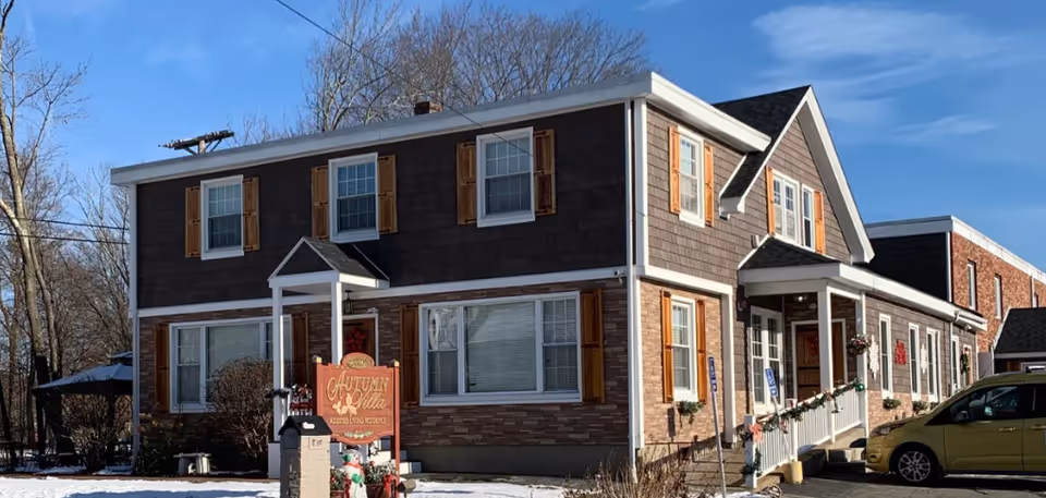 Exterior view of Autumn Villa, a two-story assisted living residence with brown siding, white trim, and wooden shutters. There is a sign in front of the building with the facility name, some holiday decorations, and a yellow car parked on the right side. The ground is covered with a light layer of snow and trees are visible in the background under a clear blue sky.