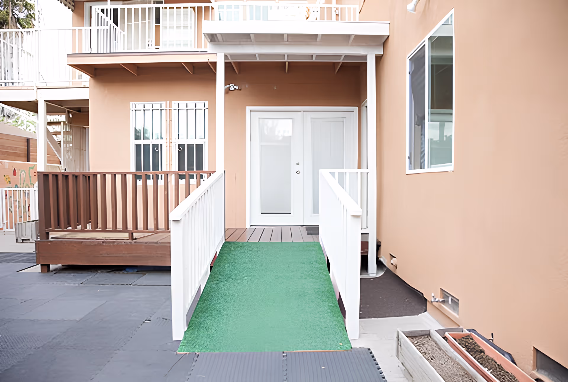 Exterior view of a building entrance with a green ramp leading up to white double doors. The building has beige walls, white railings, and a wooden deck area to the left. There is a window on the right side and some planter boxes on the ground.