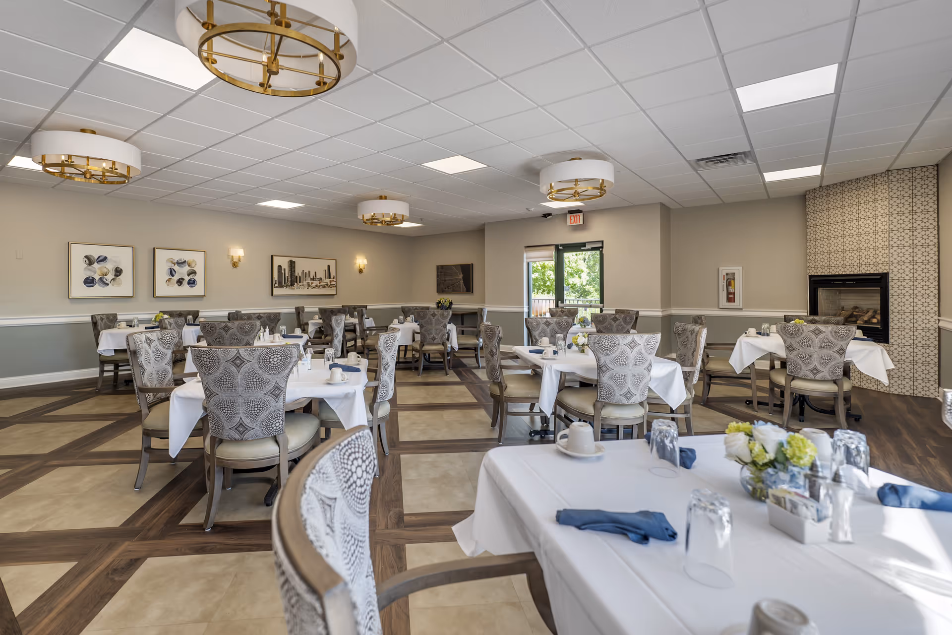 A bright and elegant dining room with multiple tables covered in white tablecloths, each set with cups, glasses, napkins, and small flower arrangements. The room features patterned upholstered chairs, modern circular ceiling lights, framed artwork on the walls, and a fireplace with a decorative tile surround. Large windows and a glass door allow natural light to enter the space.