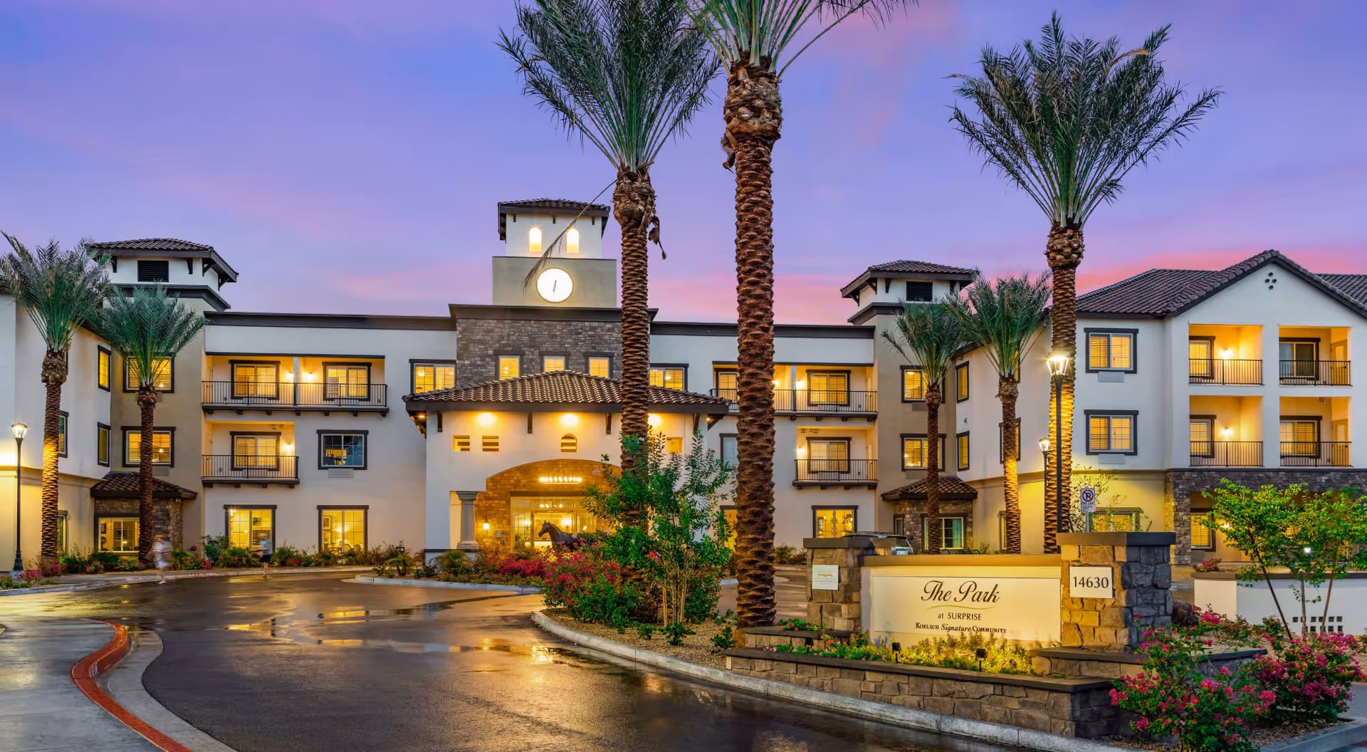 Front entrance of a three-story independent living building at dusk with palm trees, lit windows, and a stone sign reading "The Park".