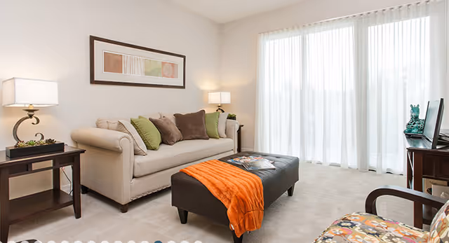 A bright living room with a beige sofa adorned with green and brown cushions, a dark brown ottoman with an orange throw blanket, a side table with a lamp, and a large window covered with sheer white curtains allowing natural light to fill the room.
