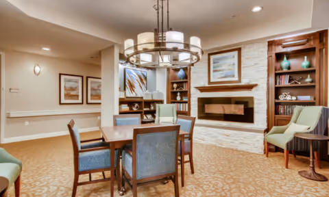 A cozy interior common area with a wooden table and six chairs in the center. The room features a modern electric fireplace set in a white stone wall, flanked by built-in wooden bookshelves with decorative items. There are framed artworks on the walls and a circular chandelier hanging above the table. The carpet has a beige pattern, and additional seating is visible along the walls.