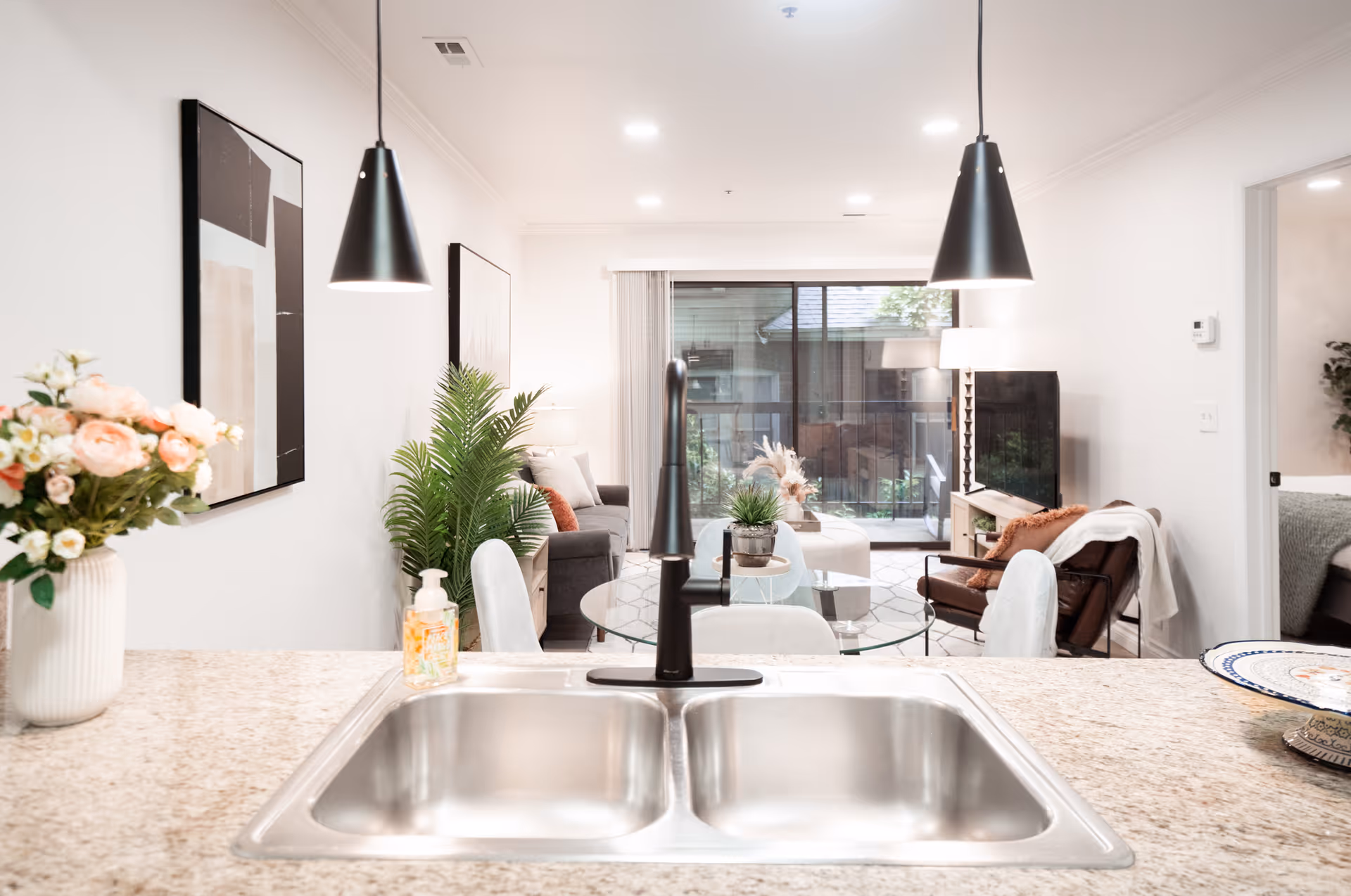 Kitchen sink and countertop facing an open living room with chairs, a couch, and sliding glass doors to a balcony.