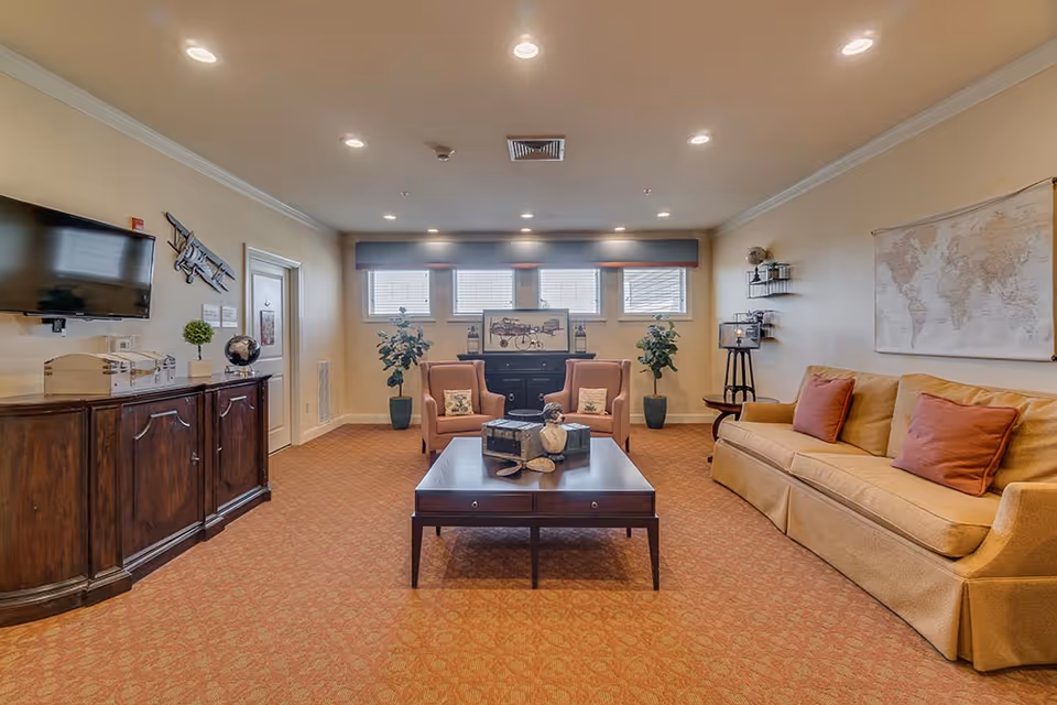 A cozy living room with a beige sofa adorned with red pillows on the right, two orange armchairs in the center facing a dark wooden coffee table with decorative items, and a dark wooden cabinet on the left with a mounted flat-screen TV above it. The room has a warm carpet, three small windows with blinds at the back, and various decorative plants and wall art including a world map and a vintage airplane model.