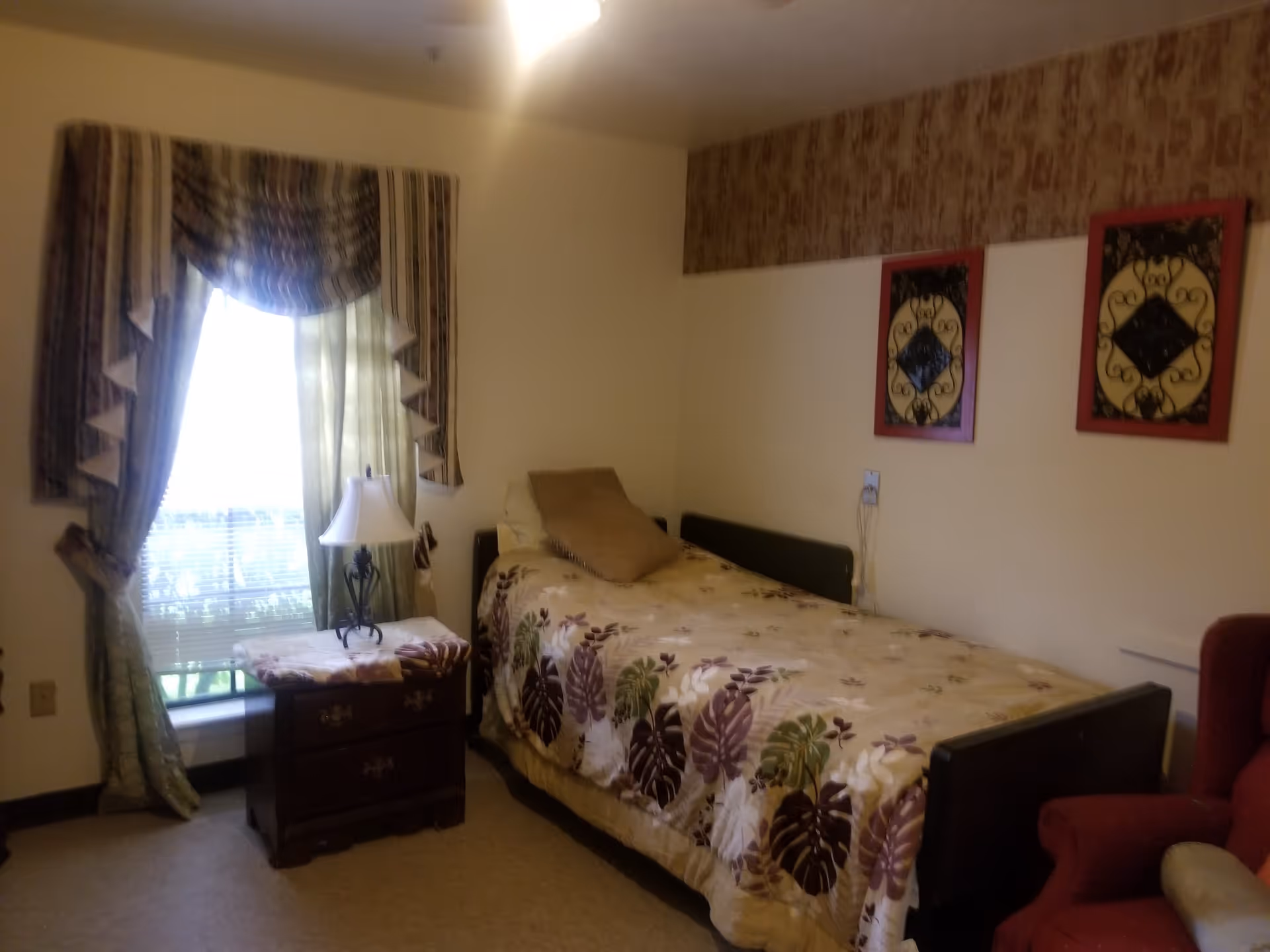 Small bedroom with a single bed covered in a floral bedspread, a nightstand and lamp by a curtained window and framed wall art.