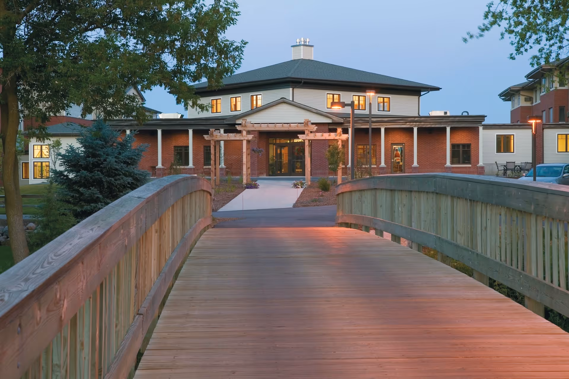 A wooden pedestrian bridge leading to the entrance of a senior living community building with brick and light-colored siding, surrounded by trees and landscaping during early evening.