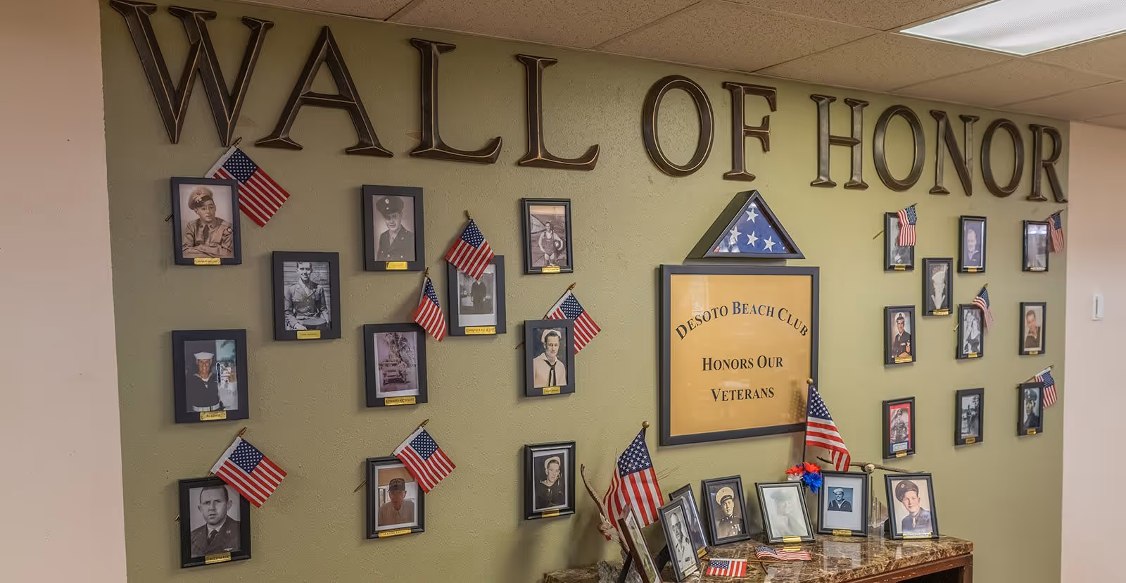 A wall display titled 'Wall of Honor' featuring framed photographs of veterans, small American flags, and a central plaque that reads 'Desoto Beach Club Honors Our Veterans'. The display is set against a green wall with a small table below holding additional framed photos and flags.