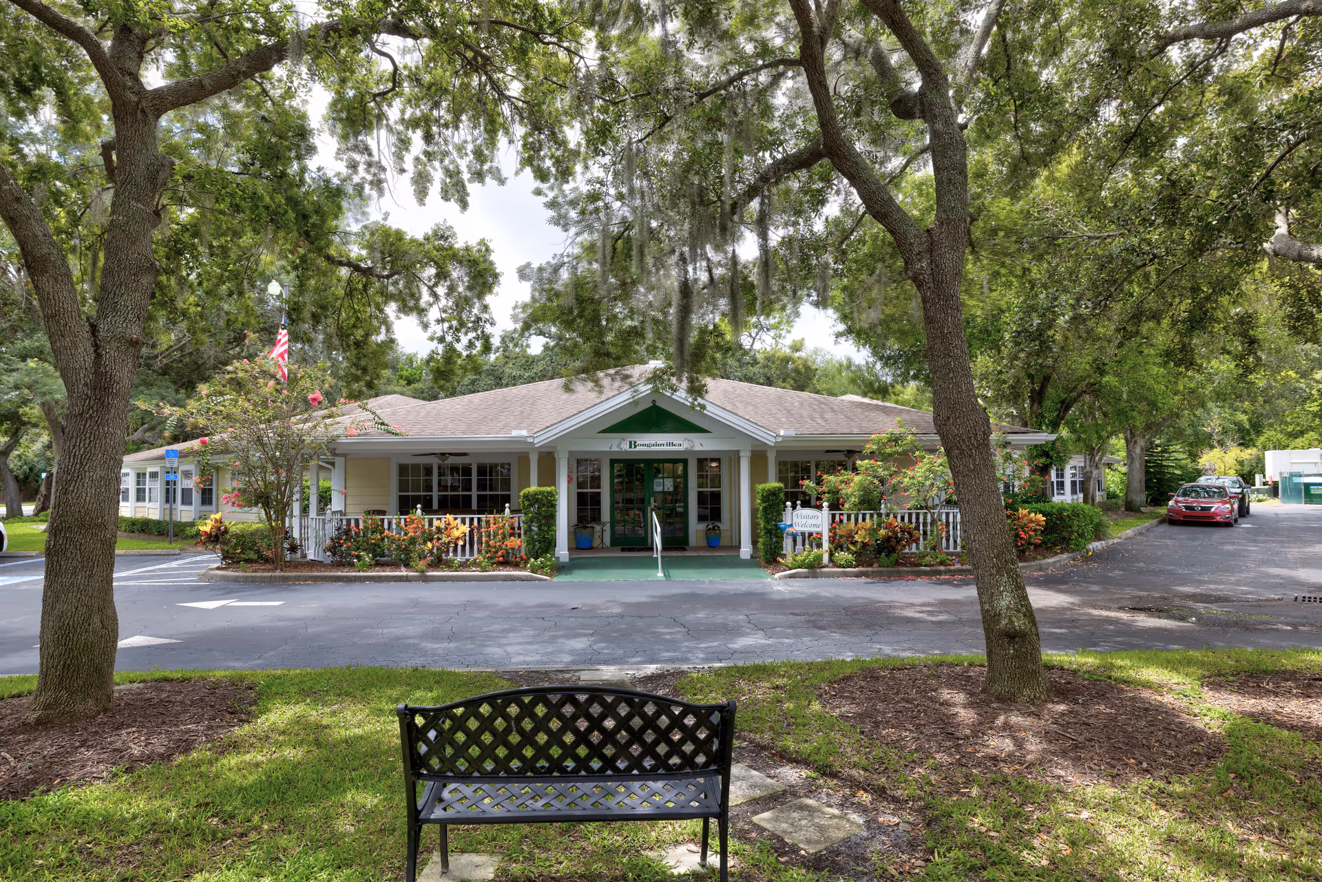 Front exterior view of a single-story building with a covered entrance, surrounded by trees and landscaping. There is a black metal bench in the foreground on a grassy area, and a parking lot with a red car to the right. The building has a sign near the entrance and an American flag is visible on the left side.