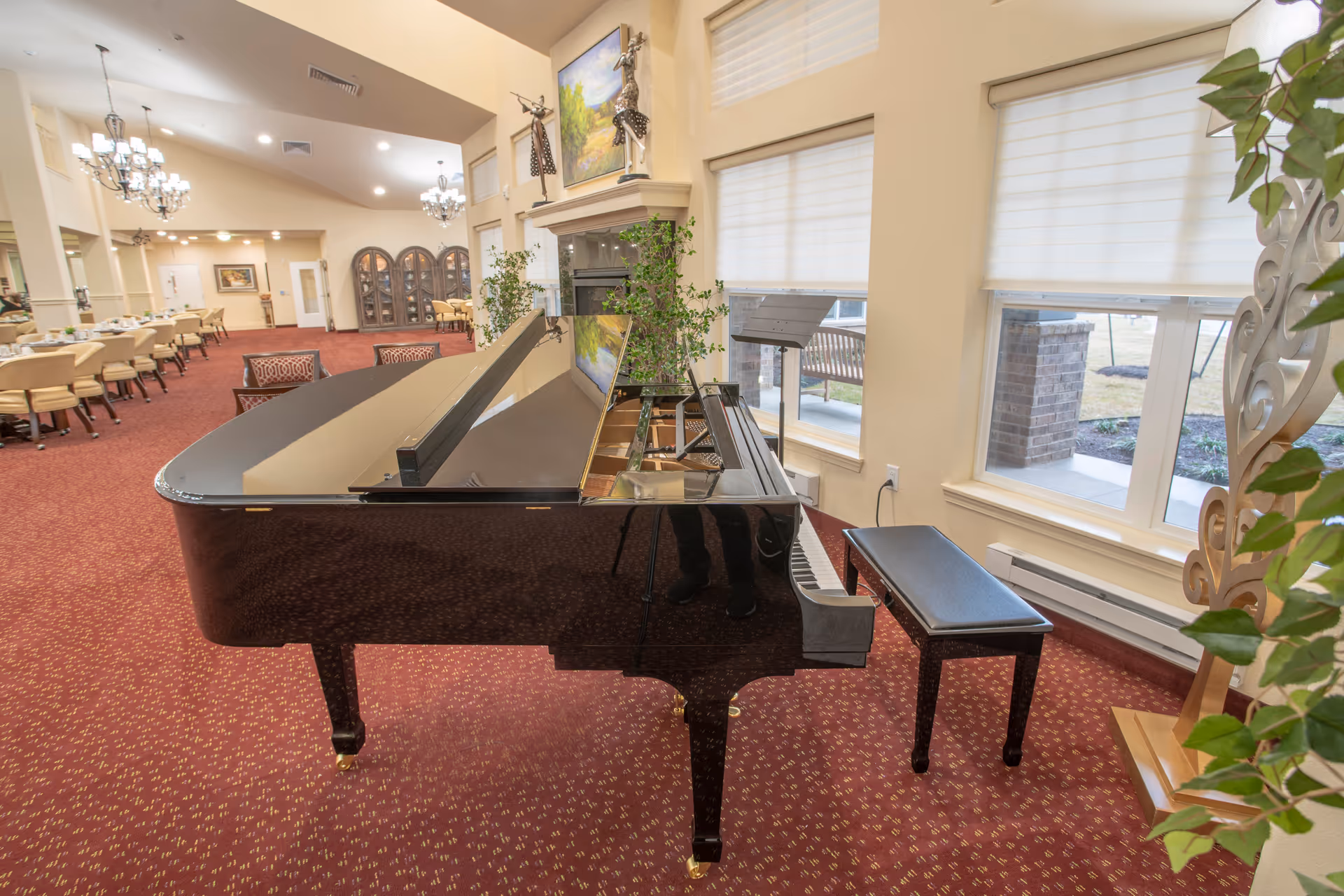 A spacious room with a grand piano in the foreground, a piano bench, large windows with blinds, and a dining area with tables and chairs in the background. The room has red carpet flooring, beige walls, chandeliers, and decorative plants.