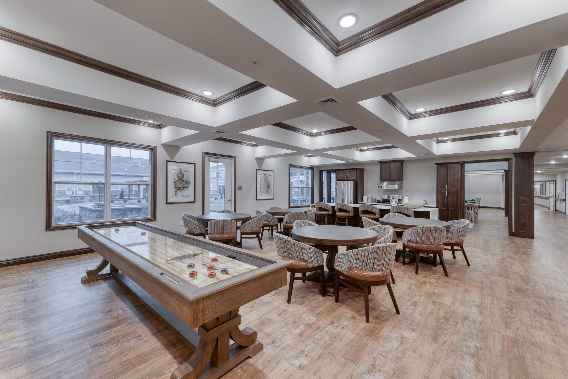 A spacious senior living common area featuring a shuffleboard table in the foreground, several round tables with cushioned chairs, and a kitchen area with stainless steel appliances and wooden cabinetry. The room has large windows, recessed lighting, and a coffered ceiling with wood trim.
