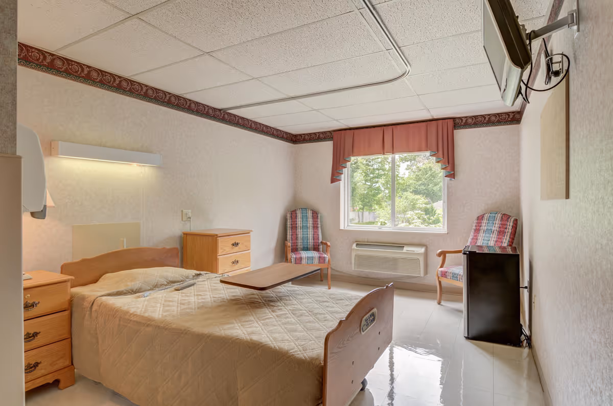 A simple, clean bedroom in a senior living facility with a bed covered in a beige quilt, two wooden nightstands, a wooden dresser, two patterned armchairs near a window with a pink valance, an air conditioning unit below the window, a small black refrigerator, and a wall-mounted television.