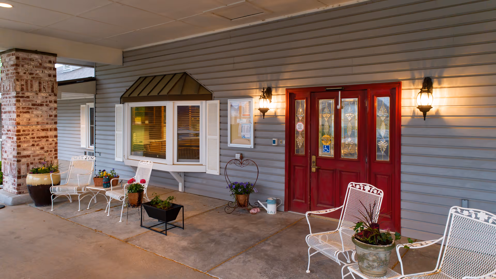 Covered outdoor entrance area of a facility with red double doors featuring decorative glass panels, two wall-mounted lantern lights, white metal chairs, and various potted plants arranged on the concrete floor and a small table.