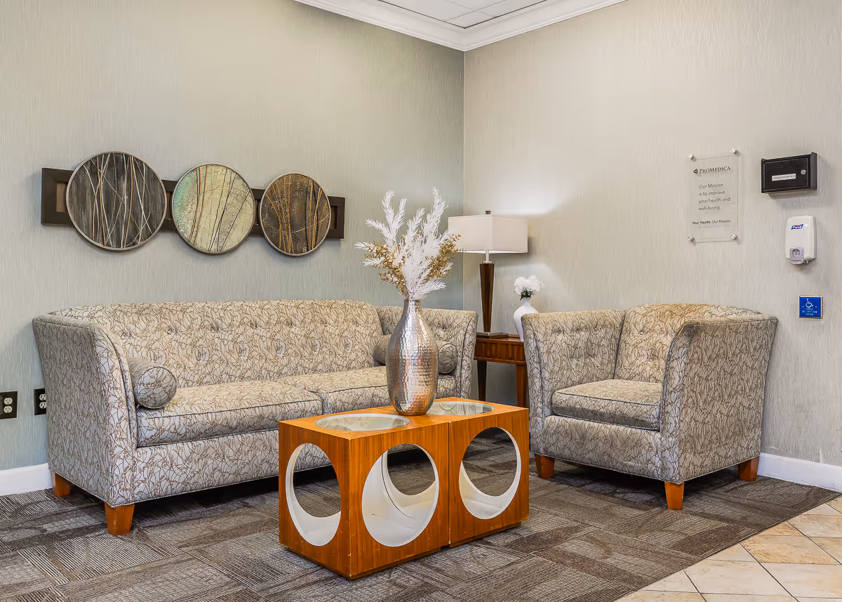 A furnished lounge with a patterned sofa and armchair, a wooden coffee table holding a silver vase, wall art, and a floor lamp.