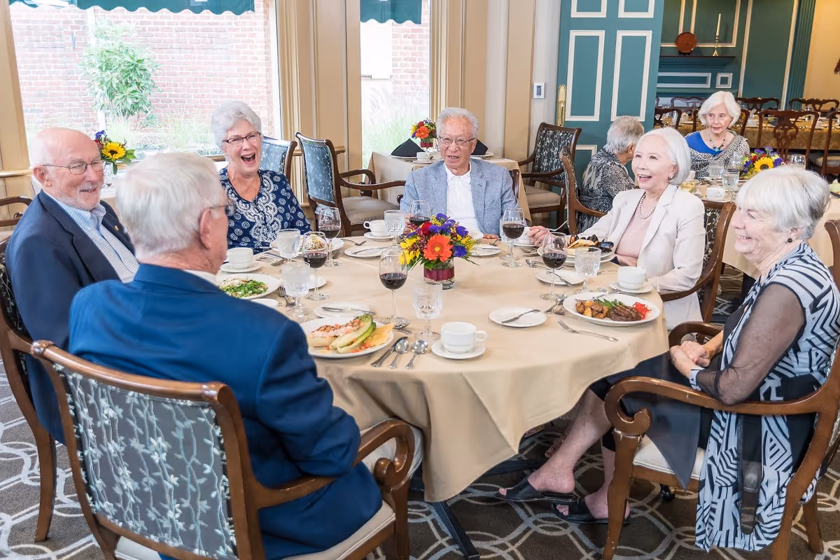 A group of elderly people sitting around a round dining table in a well-lit room, enjoying a meal together. The table is covered with a beige tablecloth and has a colorful flower arrangement in the center. Plates with food, glasses of water and wine, and cups are set on the table. The room has large windows with a view of a brick wall and greenery outside.