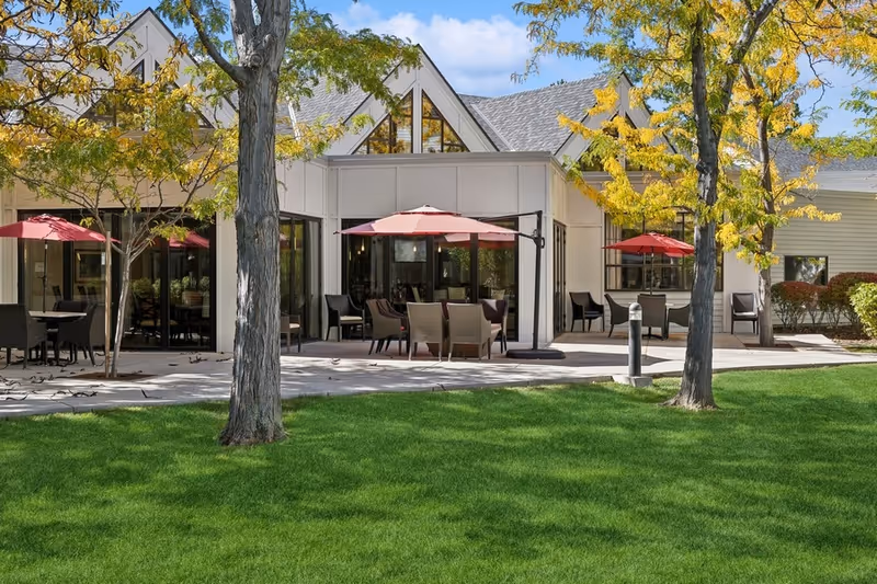 Outdoor patio area of a senior living facility with green grass, several trees with yellow leaves, and multiple seating arrangements under red umbrellas. The building has large windows and a light-colored exterior with a peaked roof.