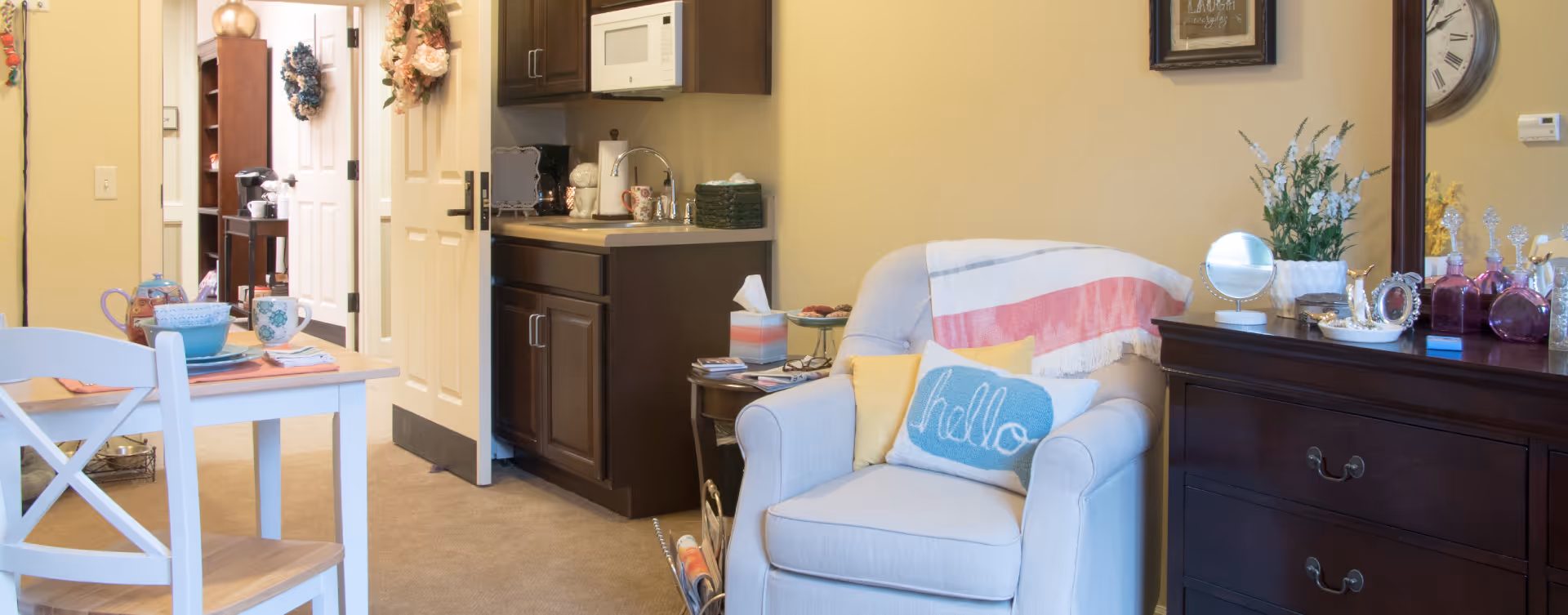 A cozy interior space featuring a small dining table set with cups and plates, a comfortable armchair with colorful pillows and a throw blanket, a dark wooden dresser with decorative items and a mirror, and a kitchenette area with dark cabinets, a microwave, and a coffee maker. The room has warm yellow walls and an open door leading to another room.