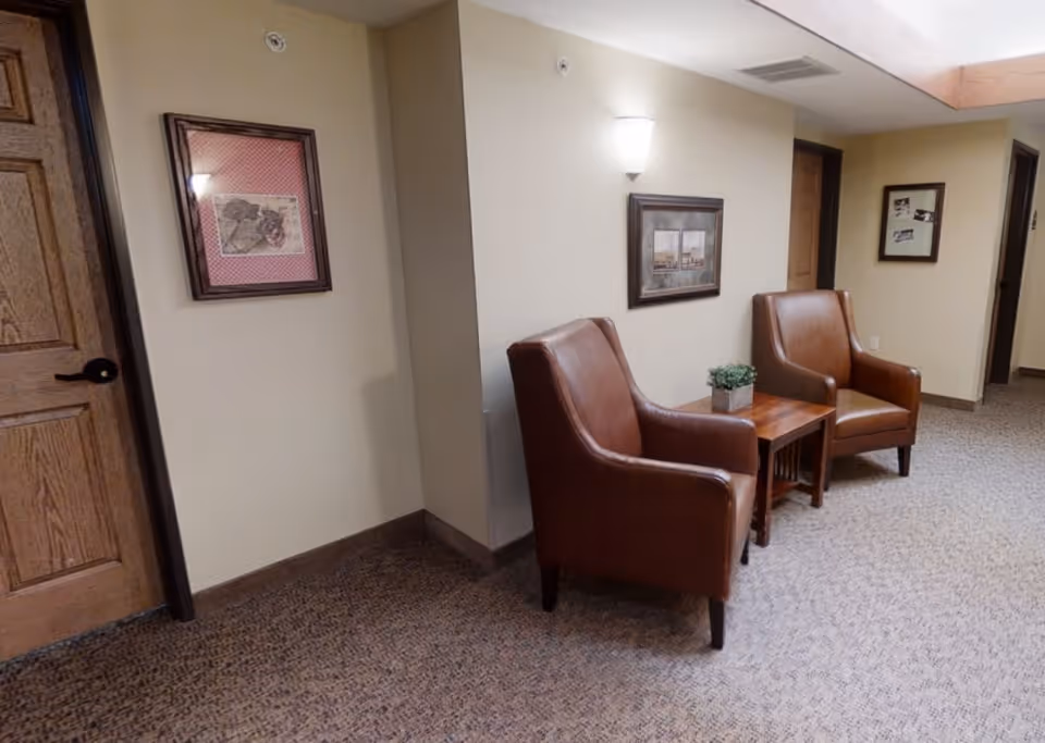 A hallway seating area in a senior living facility with two brown leather armchairs and a wooden side table with a small potted plant. The walls are beige with framed artwork, and there are wooden doors along the hallway. The floor is carpeted in a neutral pattern.
