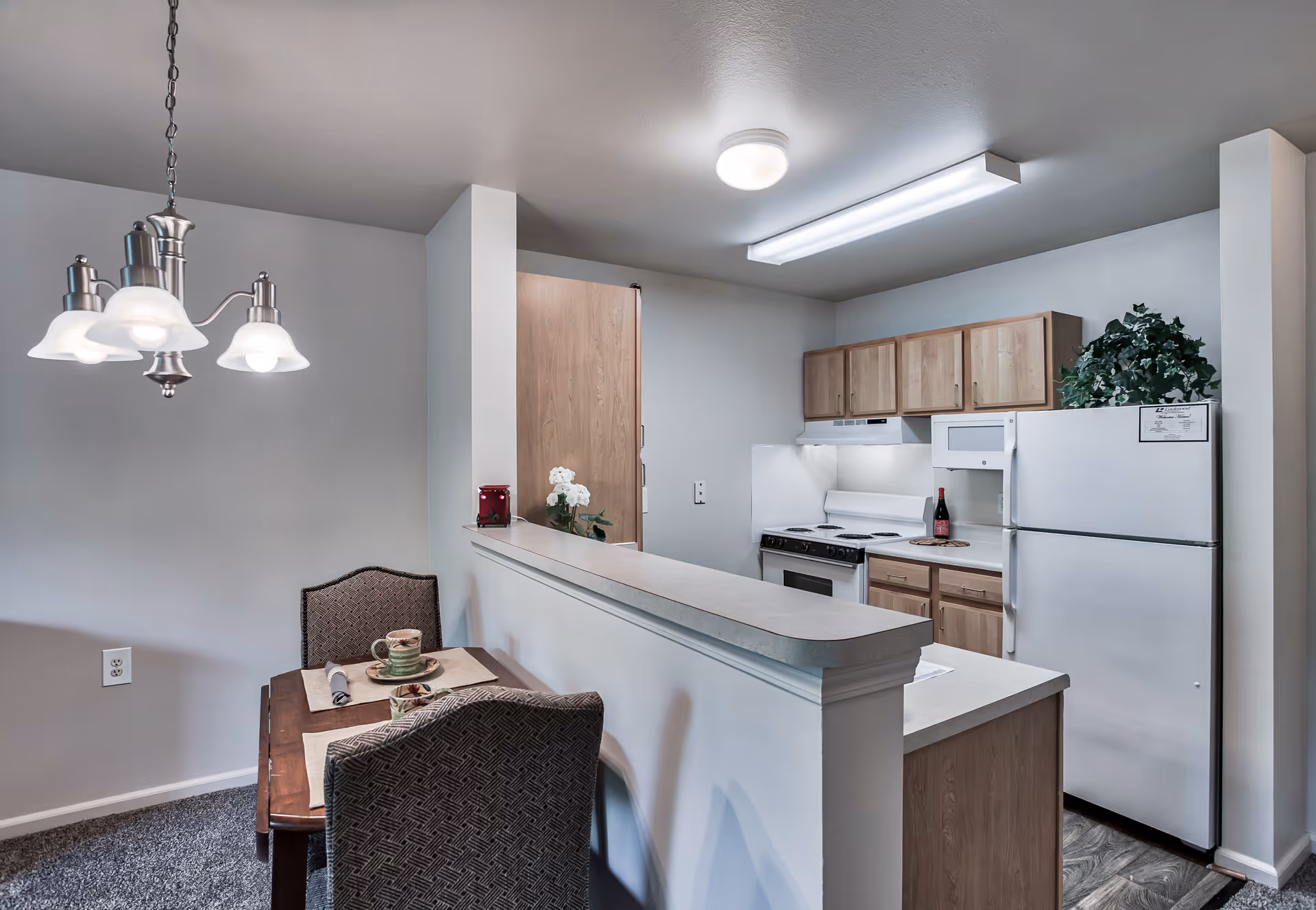 A small kitchen and dining area in a senior living facility. The kitchen features light wood cabinets, a white refrigerator, stove, microwave, and a countertop with a raised bar. The dining area has a wooden table set for two with placemats, cups, and napkins, and two upholstered chairs. A hanging light fixture is above the dining table, and there is a small plant on top of the refrigerator.