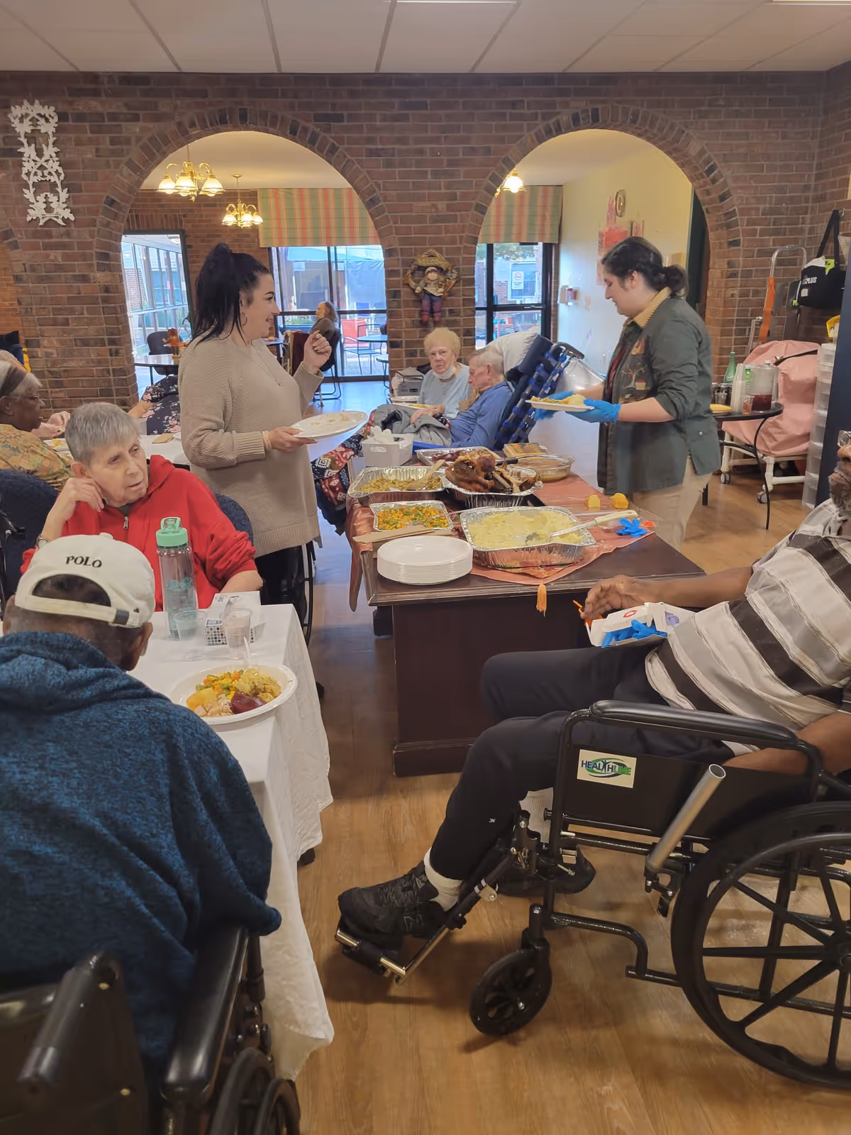 A group of elderly people and caregivers gathered in a dining area with brick archways. Several individuals are seated, some in wheelchairs, while two caregivers serve food from a table filled with trays of various dishes. The room has large windows and a warm, communal atmosphere.