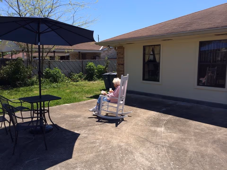 An elderly woman sitting in a white rocking chair on a concrete patio outside a building, reading a book and holding a drink. There is a black metal table with two chairs and a large black umbrella providing shade. The patio is adjacent to a grassy area with bushes and a wooden fence in the background under a clear blue sky.