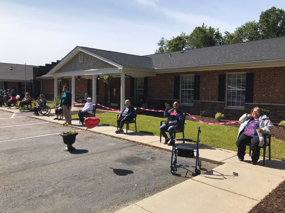Several elderly individuals sitting on chairs spaced apart outside a brick building with a covered entrance. Some people are in wheelchairs, and a walker is visible in the foreground. There is a pink ribbon stretched along the grass behind them and a heart-shaped balloon on the ground near the sidewalk.