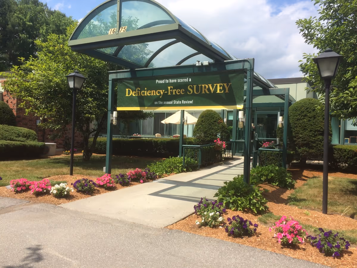Front entrance of a healthcare facility with a glass canopy, a banner reading 'Deficiency-Free SURVEY', a paved walkway and flower beds.