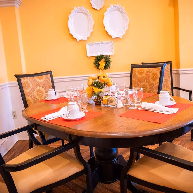 A round wooden dining table set for four with orange placemats, white cups and saucers, glassware, and napkins. The chairs have patterned cushions with a sunflower design. The background wall is painted yellow with white decorative plates and a small wreath hanging on it.
