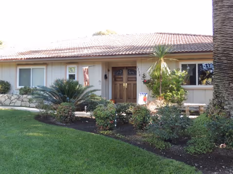 Front exterior view of a single-story building with a tiled roof, beige siding, and a wooden double door entrance. The building is surrounded by a well-maintained garden with various shrubs, plants, and a large palm tree on the right side. The lawn in front is green and neatly trimmed.