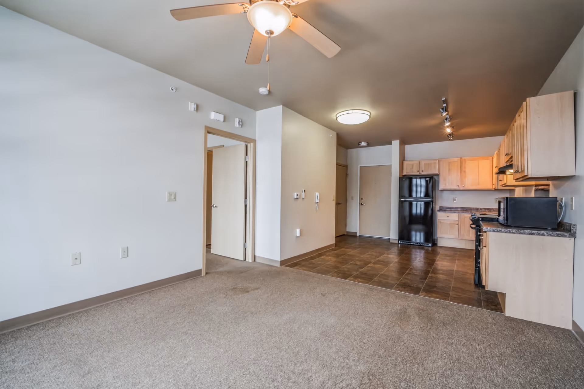 Open-plan senior apartment living area with a ceiling fan, carpeted seating area and a tile-floored kitchen with light wood cabinets and black appliances.