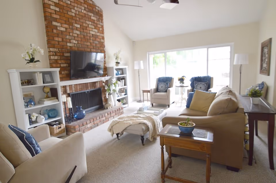 A bright living room with a brick fireplace and a mounted flat-screen TV above it. The room features beige carpeting, a beige sofa with cushions, two beige armchairs, a small wooden side table with a potted plant, and two white bookshelves flanking the fireplace. Large windows allow natural light to fill the space, and there are two floor lamps near the windows.