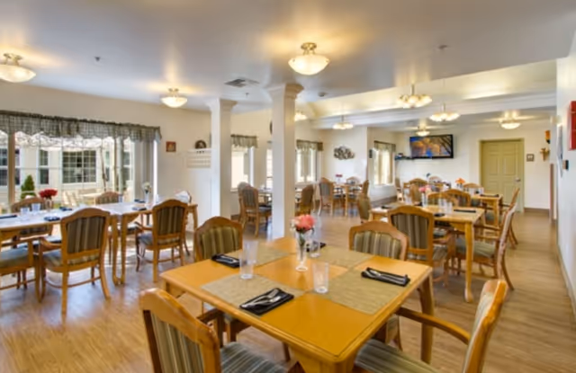 A bright and spacious dining room with multiple wooden tables and chairs arranged neatly. Each table has placemats, napkins, and glasses, with some tables decorated with small flower vases. Large windows with valances allow natural light to fill the room, and ceiling lights provide additional illumination. The room has wooden flooring and light-colored walls, creating a warm and inviting atmosphere.