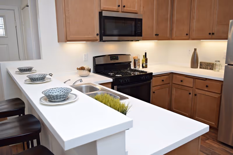Modern kitchen with wooden cabinets, a stainless steel gas stove, microwave, and refrigerator. White countertops with a double sink and three place settings with bowls and plates on the counter. Decorative items include a small plant, bottles, and a sign that reads 'My coffee needs me'.