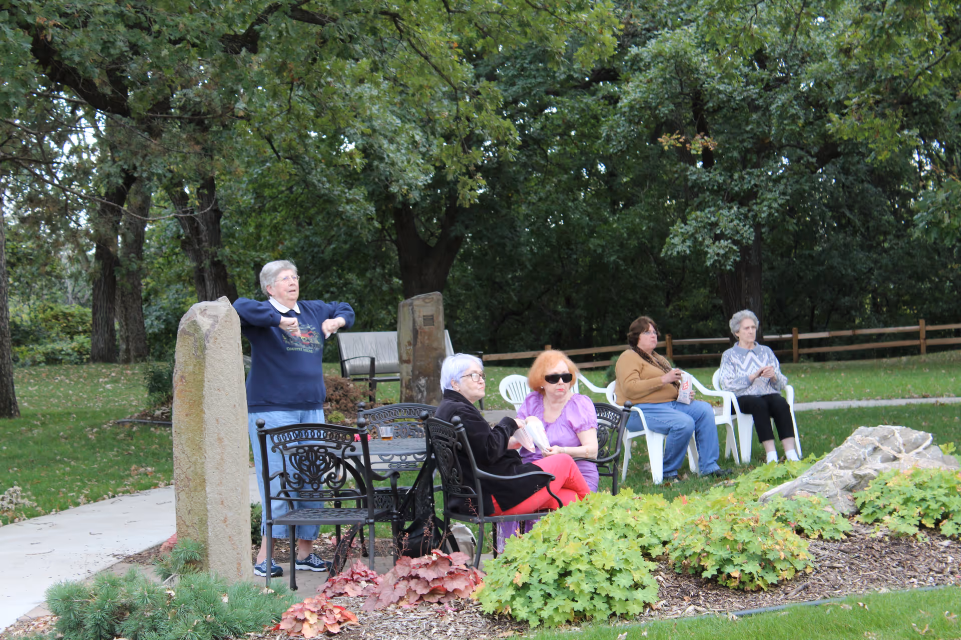 A group of elderly women sitting and standing in a garden area with green trees and plants around. Some are seated on black metal chairs and white plastic chairs, while one woman stands stretching near a stone pillar. The setting appears peaceful and outdoors.